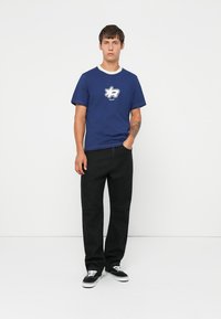 Blå kortærmet t-shirt med hvid cirkulær krave og frontgrafiklogo, parret med sorte lige ben bukser og sorte sneakers.