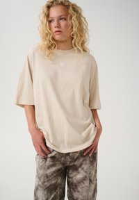 Beżowy, oversize'owy t-shirt z bawełny, z okrągłym dekoltem i krótkimi rękawami, z małym białym logo na piersi, zestawiony z szarymi spodniami w drobne plamki.