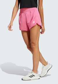 Pink athletic shorts with a gathered waistband and side slits; logo displayed on the lower left side. Worn with white sneakers.