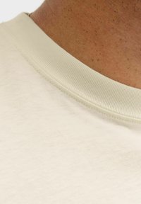T-shirt en coton beige clair avec un col rond côtelé, présentant une texture lisse et des détails de couture soignés autour du col.