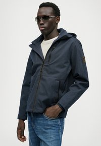 Giacca a cappuccio blu navy in tessuto texture, con zip frontale, tasche e dettagli marroni. Abbinata a un maglione chiaro e jeans di denim.