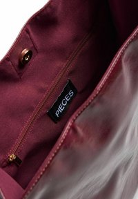 Interno di una borsa in pelle bordeaux che mostra una tasca con zip e un'etichetta nera con la scritta "PIECES" e una chiusura a scatto magnetico.