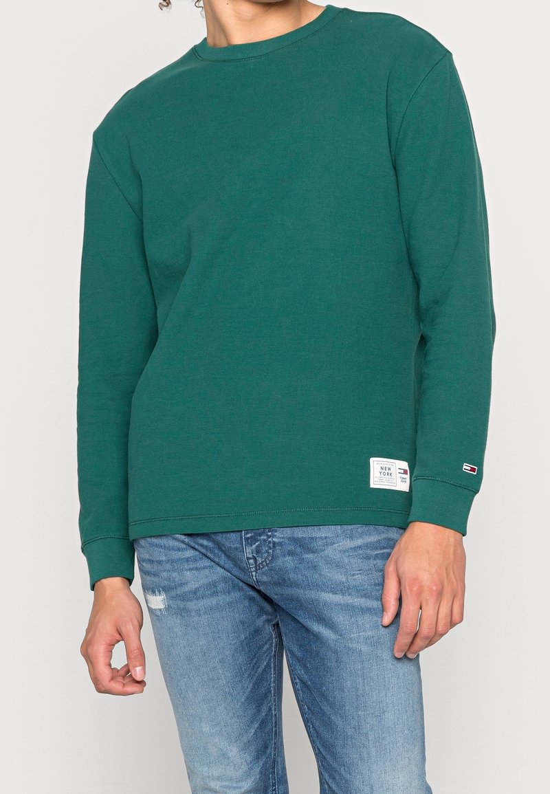 Zelená mikina s dlouhým rukávem a kulatým výstřihem vyrobená z bavlněné směsi. Obsahuje žebrované manžety a lem, s logem na spodní přední části.