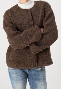 Person som bär en brun texturerad knäppbar fleecejacka och ljusblå jeans med armarna i kors, vit turtleneck synlig vid kragen.