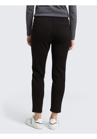 Pantaloni neri a sigaretta con vita elasticizzata, texture liscia e dettaglio della tasca posteriore, abbinati a scarpe da ginnastica bianche.