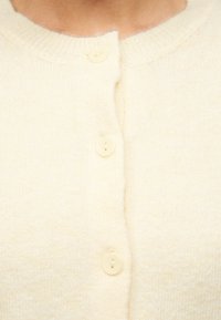 Κοντινό πλάνο ενός μαλακού, εκρού πλεκτού ζακέτου με τρία κουμπιά στο ίδιο χρώμα κλεισμένα κατά μήκος του κεντρικού μπροστινού μέρους.