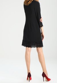 Kvinna som står med ryggen vänd, iklädd en svart klänning med spetsdetaljer och röda högklackade skor, mot en vit bakgrund.