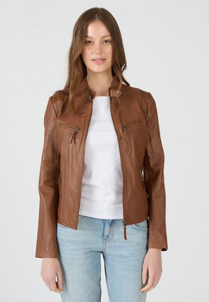 Jonge vrouw met lang bruin haar die een bruine leren jas draagt over een wit shirt en lichtblauwe spijkerbroek, staand tegen een effen achtergrond.