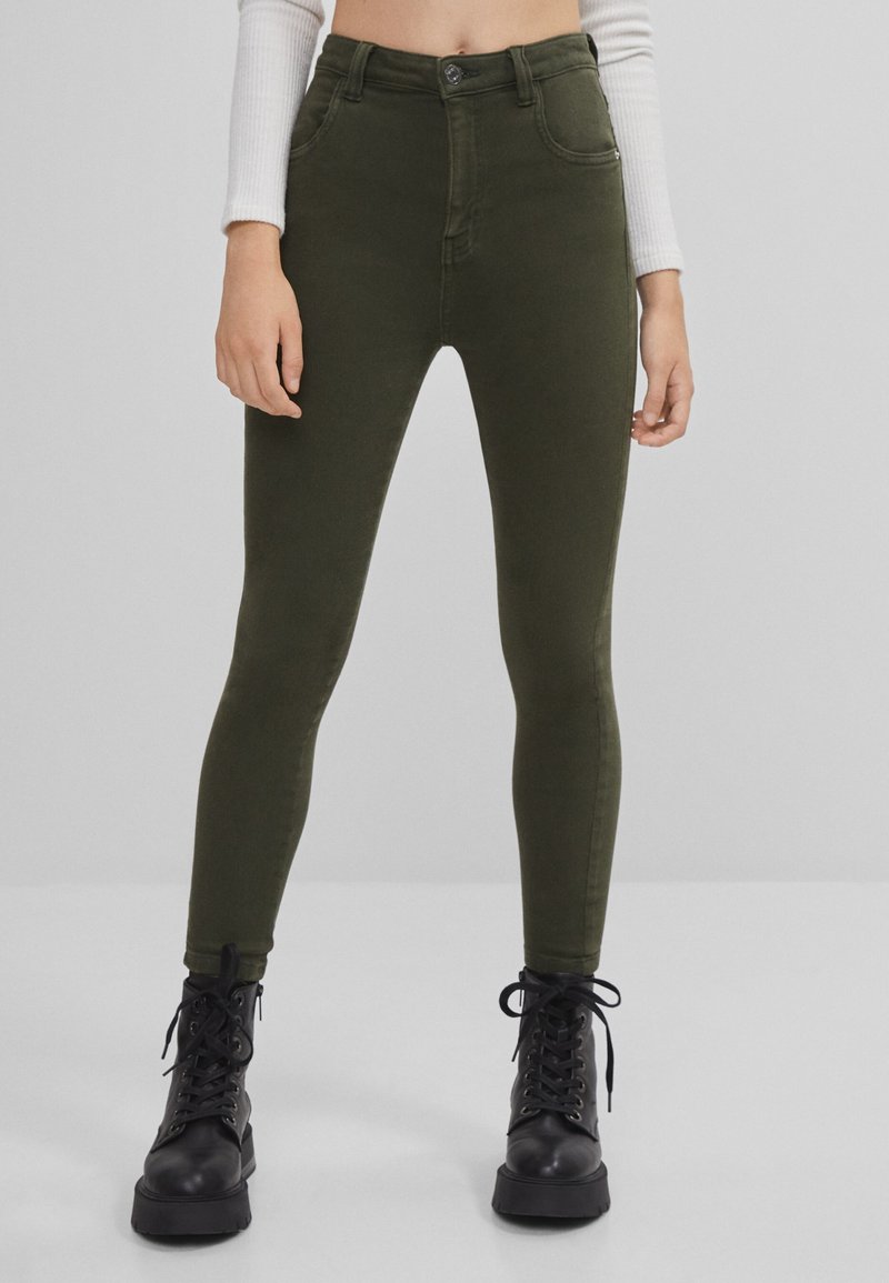 Femme portant un jean skinny taille haute couleur olive avec poches avant et bottines noires à lacets, sur un fond gris clair uni.