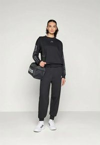 Melna sporta apģērba komplekts, kas sastāv no garām piedurknēm un šaurām biksēm, abiem ir smalki logotipa akcenti, kopā ar melnu somu un baltām kurpēm.