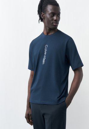 Homme à la peau foncée et aux dreadlocks portant un t-shirt bleu marine Calvin Klein avec un logo vertical, debout avec une main dans la poche, regardant sur le côté.