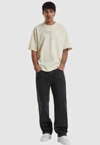 T-shirt en coton oversize jaune clair à manches courtes avec un design brodé, associé à un pantalon en denim noir et des baskets blanches.
