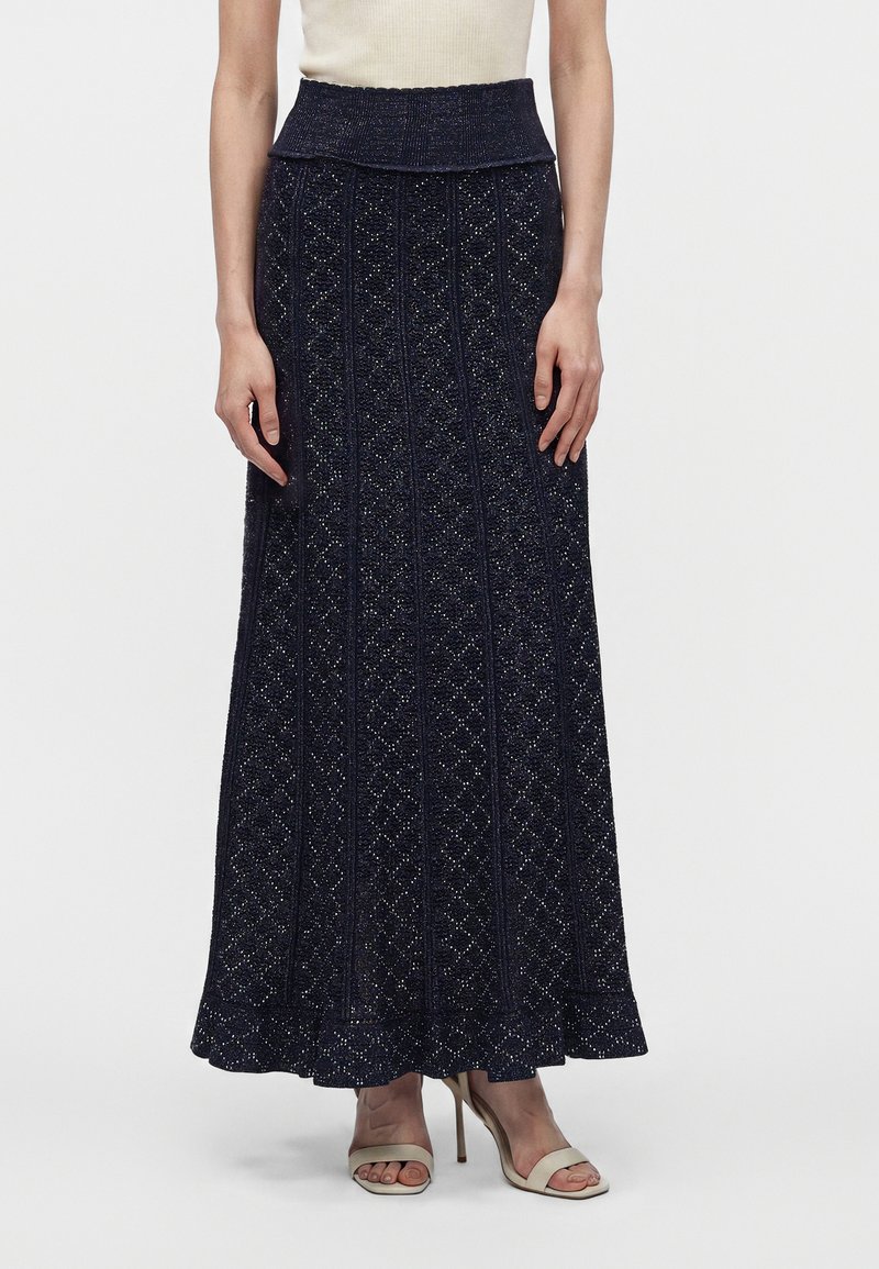 Femme portant un haut sans manches crème, une jupe en maille bleu marine longueur cheville à motif texturé, et des sandales à talons hauts beiges.