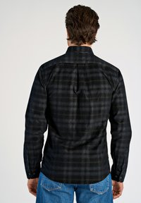 Man gezien van achteren in een zwart-grijs geruite lange mouw shirt en blauwe spijkerbroek tegen een effen achtergrond.