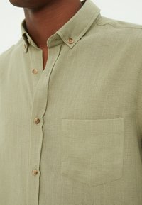 Camisa de botones verde claro con una tela texturizada, que presenta un collar, botones de madera y un bolsillo en el pecho.