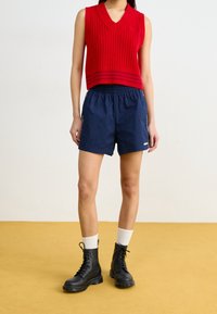 Frau in einem roten ärmellosen Stricktop, dunkelblauen Shorts mit elastischem Bund, weißen Socken und schwarzen Schnürstiefeln, die auf einem senfgelben Boden steht.