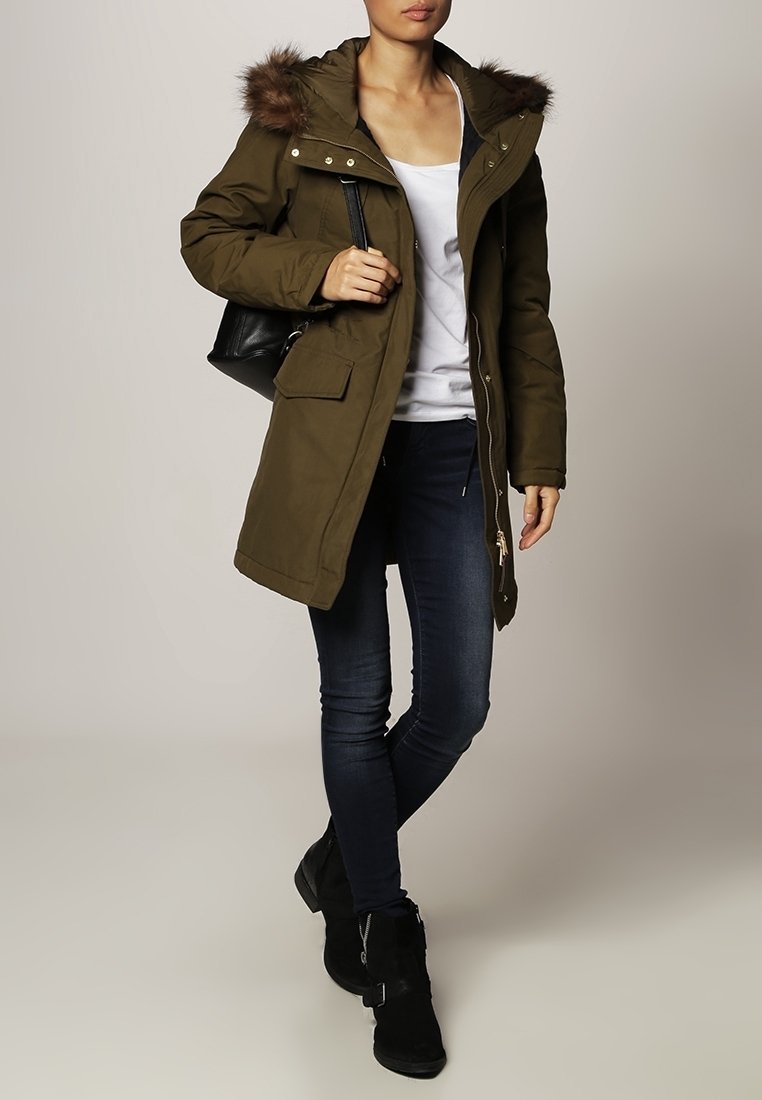 Parka rembourrée vert olive avec capuche bordée de fourrure, deux poches avant, fermetures éclair, et coupe décontractée, associée à un jean foncé et des bottines.