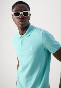 Polo shirt turquoise au design ajusté, avec deux boutons au col et un logo brodé discret sur la poitrine. Porté avec des lunettes de soleil blanches.