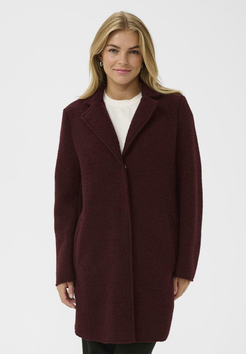 Burgundska volnena plašč s izrezanim ovratnikom, enojnim gumbom, dvema stranskima žepoma in dolgimi rokavi. Teksturiirana tkanina z mehak videzom.