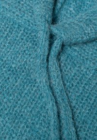 Zbliżenie na miękki, teksturowany materiał w kolorze zielononiebieskim, z widocznym splotem i szczegółem w postaci węzła na środku.