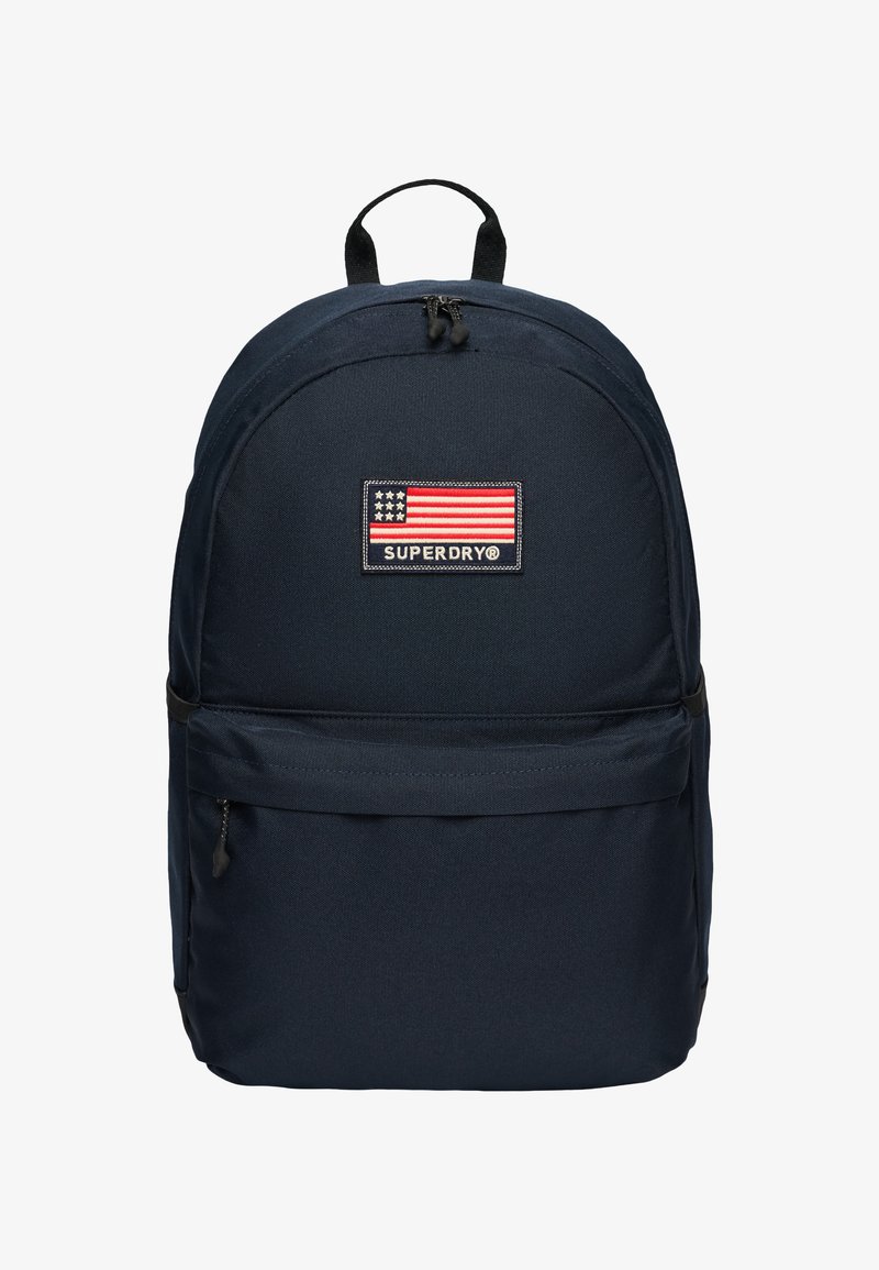 Zaino blu navy con tasca frontale con zip, manico superiore e patch con logo Superdry che presenta una bandiera americana stilizzata.