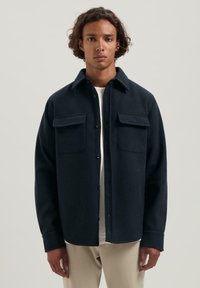 Granatowa koszula zapinana na guziki z kołnierzykiem, dwiema kieszeniami na piersi i miękkim, teksturowanym materiałem. Posiada luźny krój i długie rękawy.