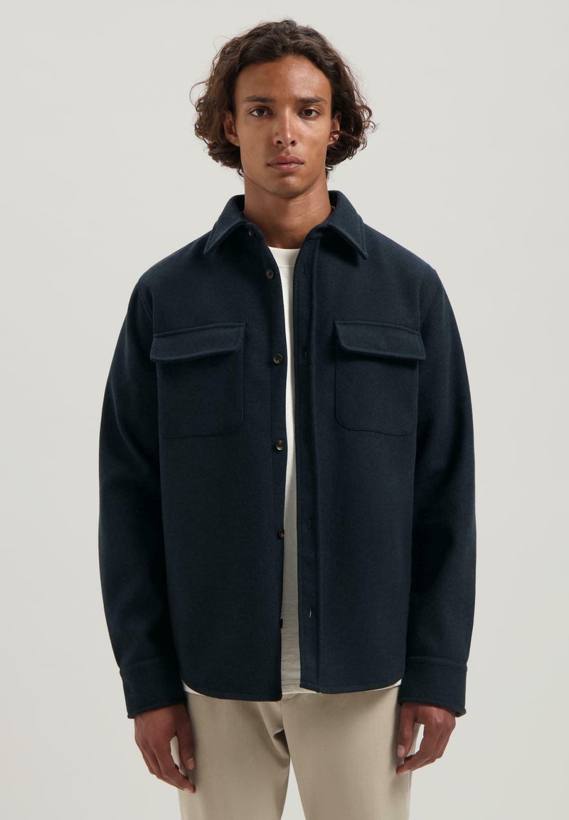 Granatowa koszula zapinana na guziki z kołnierzykiem, dwiema kieszeniami na piersi i miękkim, teksturowanym materiałem. Posiada luźny krój i długie rękawy.