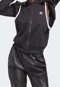 Schwarze Zip-Jacke mit Kapuze, ausgestattet mit weißen Akzenten, vorderen Taschen und einem kleinen Adidas-Logo. Kombiniert mit glänzenden schwarzen Hosen.