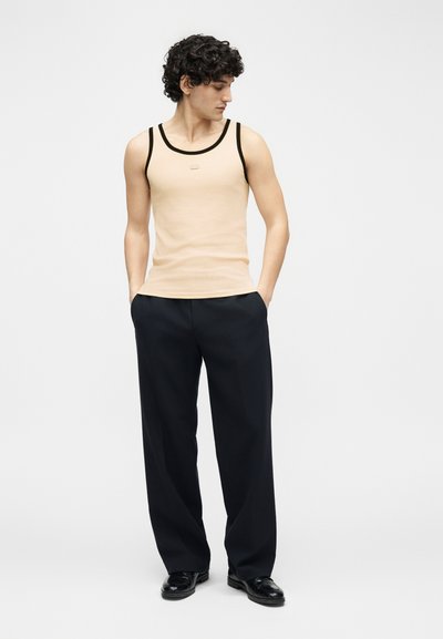 Jeune homme aux cheveux bouclés portant un débardeur beige avec des bordures noires, un pantalon large noir et des chaussures noires, debout les mains dans les poches.