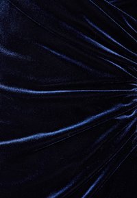 Tmavě modrý sametový látka s měkkými, nepravidelnými záhyby vytvářejícími texturovaný, lesklý povrch odrážející světlo.