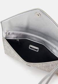 Pochette pailletée argentée ouverte montrant l'intérieur en tissu noir avec une poche zippée et une petite étiquette de marque en métal à l'intérieur du rabat.