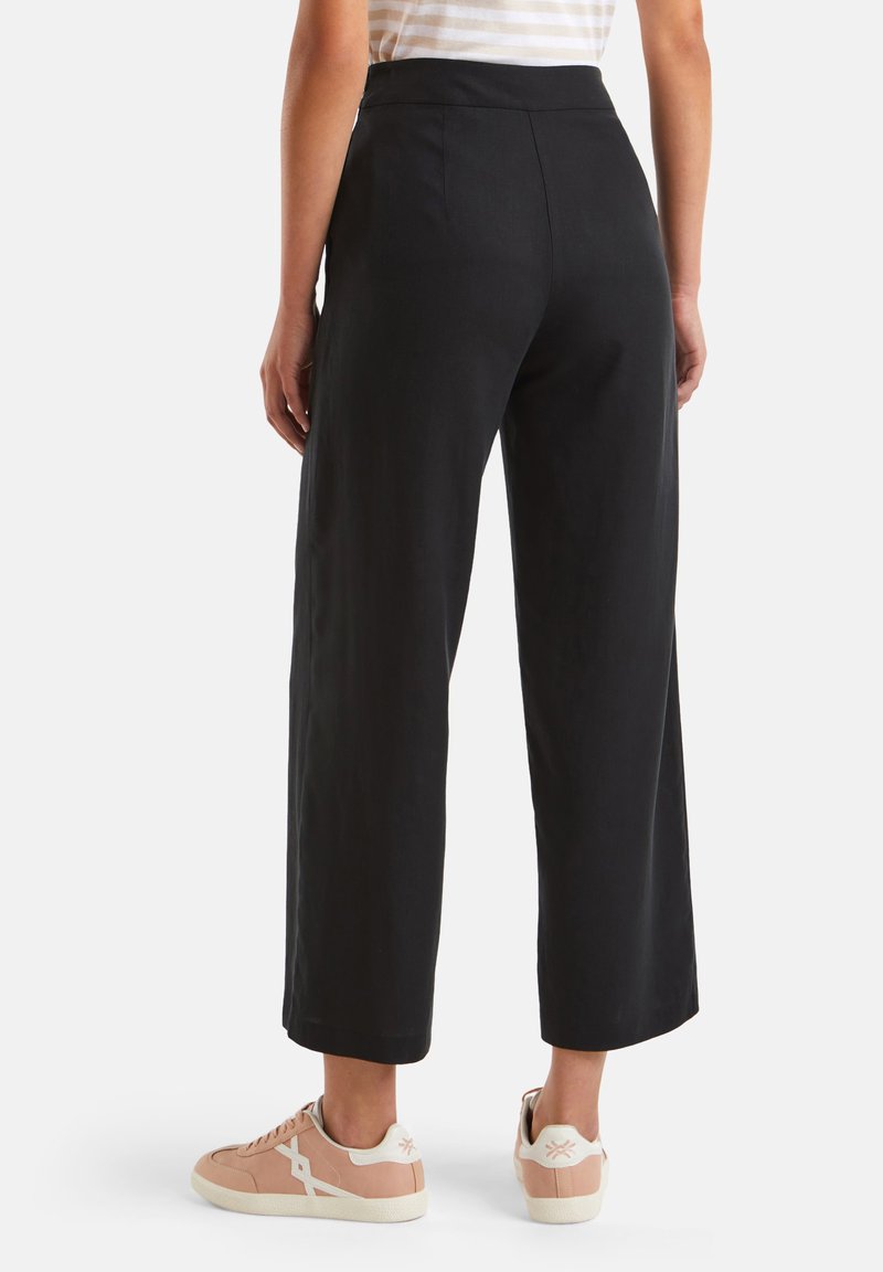 Schwarze weite Hose aus glattem Material, mit mittelhohem Bund und seitlichen Taschen. Kombiniert mit hellrosafarbenen Sneakern mit weißen Akzenten.