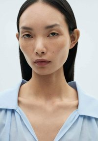 Camicia azzurra chiaro con colletto in un tessuto morbido e trasparente. Il modello ha la pelle liscia, capelli neri lisci e un trucco minimal. Nessun motivo o accessorio.