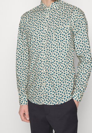 Mężczyzna ubrany w kremową, długorękawową koszulę na guziki z małym niebieskim kwiatowym wzorem oraz czarne spodnie, stojący z jedną ręką w kieszeni.