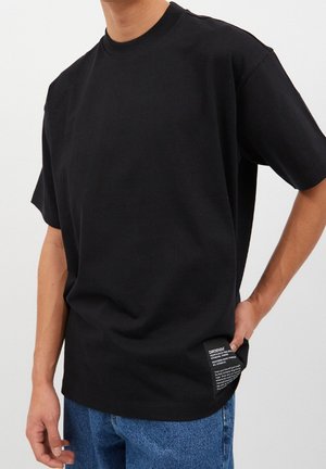 Mężczyzna ubrany w czarny, gładki  T-shirt z krótkim rękawem, z białą metką z napisem przy dolnym brzegu i niebieskie dżinsy, ręka w kieszeni na białym tle.