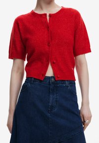 Punainen lyhythihainen neuletakki, jossa on pyöreä kaulus ja edessä napit. Pehmeä materiaali, lyhyt muotoilu, yhdistettynä tummansinisiin farkkuhameeseen.