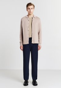 Hellbeige Strickjacke mit Knöpfen und Kragen, über einem taillierten hellbraunen Hemd getragen, kombiniert mit navyblauen, maßgeschneiderten Hosen und schwarzen Schuhen.