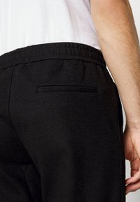 Černé tepláky mají strukturovanou tkaninu, elastický pas a jedno zadní kapsu. Důraz je kladen na zadní pohled na oděv.