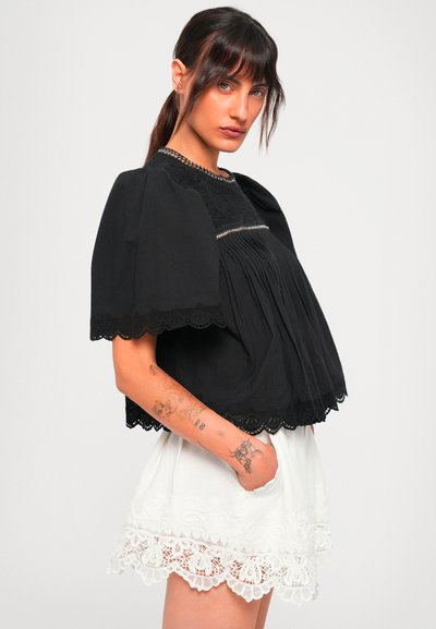 Woman with tattooed arm wearing a black pleated blouse with puff sleeves and white lace-trimmed shorts, standing against a plain background.
