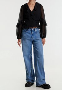 Haut cache-cœur noir à volants en tissu texturé, associé à un jean bleu clair à jambes larges et des bottes noires épaisses.