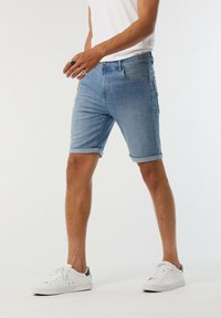 Shorts en denim bleu avec des revers roulés, présentant un design classique à cinq poches et une coupe slim. Portés avec des baskets blanches et une chemise unie.