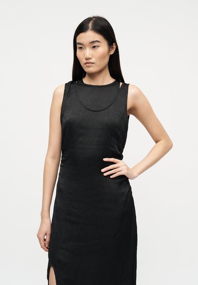 Νεαρή γυναίκα με μακριά σκούρα μαλλιά, φορώντας αμάνικο μαύρο φόρεμα με υφή, στέκεται με το χέρι στη μέση μπροστά σε απλό λευκό φόντο.