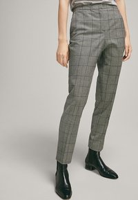 Pantalon à carreaux gris avec une coupe ajustée, taille haute, devant plat et jambe slim. Associé à des bottines noires pour un look soigné.