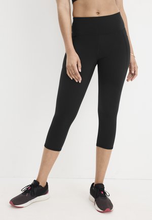 Mujer vistiendo leggings negros cortos con bolsillos laterales y zapatillas negras para correr sobre un fondo blanco liso.