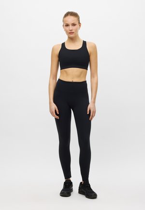 Frau steht, trägt einen schwarzen Sport-BH, hoch taillierte Leggings und schwarze Sneakers vor einem einfarbigen hellen Hintergrund.
