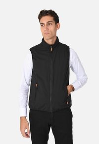 Gilet senza maniche nero in tessuto leggero, caratterizzato da un colletto alto, zip frontale e tirazip arancioni. Indossato sopra una camicia bianca a maniche lunghe.