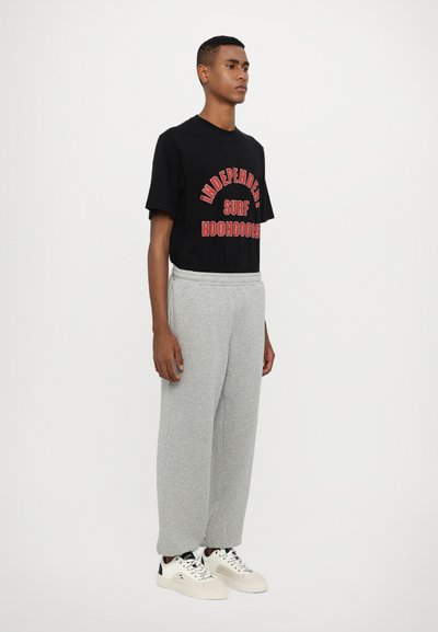 Black t-shirt with red graphic text, paired with light gray sweatpants. Model wears white sneakers with black accents. Plain background.