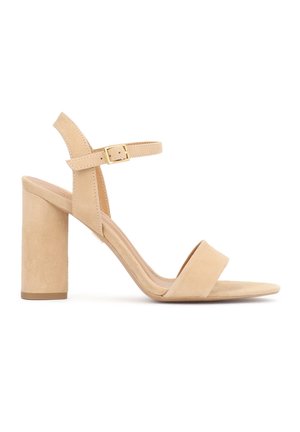 Smėlio spalvos zomšiniai aukštakulniai basutės su ankleidžiama diržele, auksine sagtimi, atvira nosimi ir storu kulnu ant baltos spalvos fono.