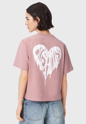 Person med kort brunt hår har en lyserød t-shirt med et hvidt, smeltende hjertemotiv og blå jeans på, set bagfra.