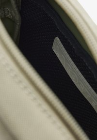 Bolsa de tela beige con una superficie texturizada, cuenta con un interior negro, cierre de cremallera y una etiqueta translúcida en el interior.